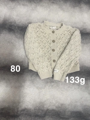 Beige stickad kofta från Newbie - Säljer en beige stickad kofta från Newbie med snyggt hålmönster och ribbade muddar. Koftan har rund hals och knäpps med fem bruna knappar framtill. Perfekt för lager-på-lager och mysiga dagar.🌸 Fint använt skick. Storlek 80