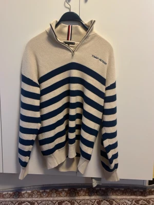 Randig stickad tröja Tommy Hilfiger - Säljer en snygg stickad tröja från Tommy Hilfiger i storlek 164. Tröjan är beige med mörkblå horisontella ränder, har en half zip-dragkedja vid kragen och långärmade ribbade muddar. Perfekt för dig som gillar klassisk stil med en modern touch.