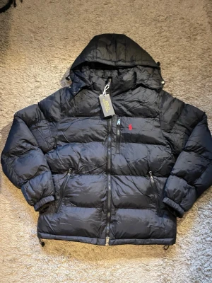 Svart pufferjacka från Polo Ralph Lauren - Svart pufferjacka från Polo Ralph Lauren med huva, två sidofickor med dragkedja och en röd broderad logga på bröstet. Jackan har quiltad design och hel dragkedja framtill. Perfekt för kalla dagar och har en riktigt chill vibe.