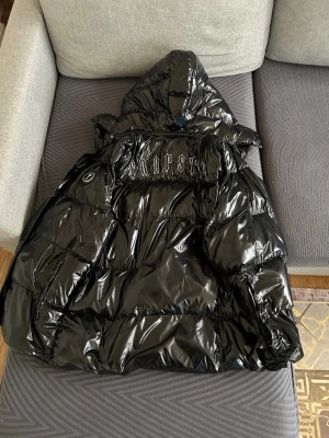 Svart dunväst från Trapstar London - Säljer en svart, glansig dunväst från Trapstar London med huva och broderad logga på ryggen. Västen har dragkedja och tryckknappar framtill samt stora fickor. Perfekt för lager-på-lager och streetwear-stil.