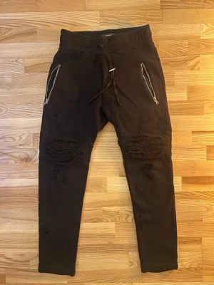 Svarta Amiri mjukisbyxor med dragkedjor - Svarta mjukisbyxor från Amiri i 100% bomull med detaljer i äkta läder. Byxorna har dragkedjor vid fickorna, snörning i midjan och slitna partier över knäna för en edgy look. Tight passform och ribbade detaljer ovanför knäna.