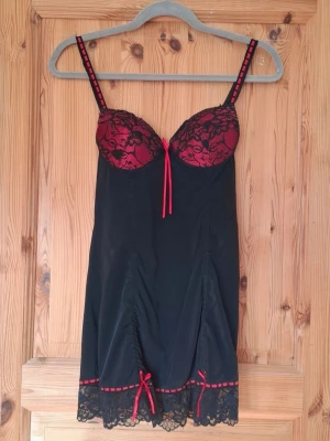 Svart och röd negligé med rosetter - Supersöt svart neglige med röda detaljer. Spetsdetaljer, rosetter och justerbara axelband. Stretchigt material. Sparsamt använd men fortfarande i fint skick. Jag har inte hittat något att anmärka på. B75 kupa. 90% nylon, 10% spandex.