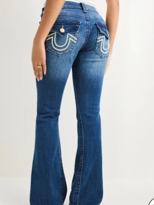 Blå bootcut true religion jeans med broderade fickor - Snygga blå jeans med bootcut-modell och broderade bakfickor med lock och knapp. Jeansen har en klassisk femficksdesign och markerade sömmar som ger en cool look. Perfekta för dig som gillar en trendig och avslappnad stil. Lite slitna nere vid bena