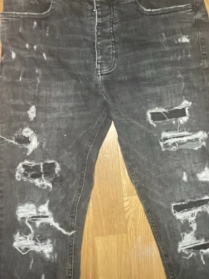 Svarta ripped jeans från Valére Milano - Säljer ett par svarta jeans från Valére Milano med snygga slitningar och hål på framsidan för en edgy look. Jeansen har klassisk femficksdesign och är tillverkade i jeansmaterial med en mörk tvätt. Passformen är rak och de har normal midja.