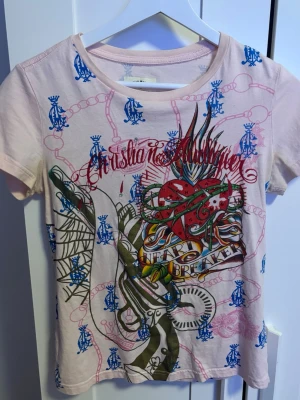 Christian Audigier rosa t-shirt XL - Unik rosa t-shirt från Christian Audigier med färgstarkt tryck fram och bak, inklusive hjärta, ankare och text. T-shirten har korta ärmar, blå och gula loggor samt detaljer i rött och grönt. Tillverkad i 100% bomull. Står att den är i storlek XL men passar mer som M/S