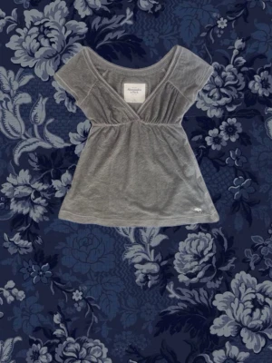 Grå omlottopp från Abercrombie & Fitch - Supersöt grå topp från Abercrombie & Fitch med omlottdesign och korta ärmar. Toppen har en v-ringad skärning och är tillverkad i mjuk bomull, perfekt för en chill och stilren look.