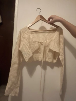 Beige stickad croppad kofta med knyt - Superfin beige stickad kofta med croppad passform och långa ärmar. Koftan har en snygg knytning framtill och är perfekt att slänga över ett linne eller klänning. Mjuk och mysig, passar perfekt för lager-på-lager-stil.
