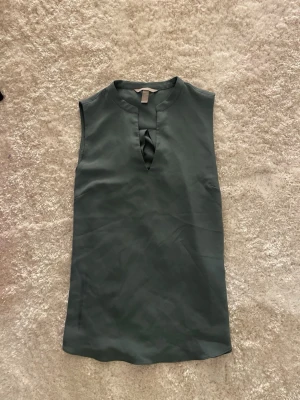 Grön ärmlös blus från H&M - Snygg mörkgrön ärmlös blus från H&M i storlek 36. Blusen har en stilren v-ringning med liten twistdetalj framtill och en lätt A-linjeformad siluett. Perfekt att matcha med jeans eller kjol för en clean look.