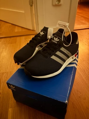 Adidas LA Trainer sneakers svart/silver - Säljer ett par Adidas LA Trainer sneakers i svart med silvriga ränder och detaljer. Skorna har snörning, rund tå och en platt sula med gummidubbar undertill. Ovandelen är i syntetmaterial och mesh för bra ventilation. Klassisk Adidas-logga på plösen och sidan.