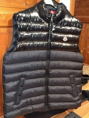 Svart dunväst från Moncler - Säljer en svart dunväst från Moncler med glansig finish upptill och matt nedtill. Västen har ståkrage, två fickor med knappstängning och dragkedja framtill. Insidan är röd med coola Moncler-etiketter och innerficka med dragkedja. Perfekt för dig som gillar stil och funktion.