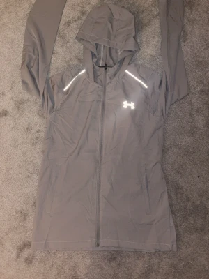 Grå vindjacka från Under Armour - Snygg grå vindjacka från Under Armour med huva och hel dragkedja framtill. Jackan har reflekterande detaljer på axlarna och logga på bröstet samt stor logga på ryggen. Tillverkad i lätt polyester, perfekt för träning utomhus.