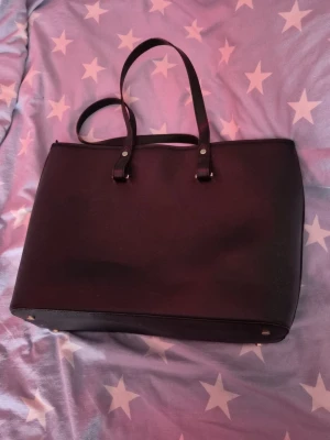 Svart handväska från H&M - Stilren svart handväska från H&M med två långa handtag och dragkedja upptill. Väskan har en enkel, klassisk design och är tillverkad i syntetmaterial. Insidan har ett fack med dragkedja för småsaker. Perfekt för dig som vill ha plats för allt du behöver under dagen.