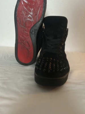 Svarta Louboutin sneakers med nitar - Säljer ett par svarta high-top sneakers från Louboutin i mocka med ikonisk röd sula och coola svarta nitar över tån. Skorna har snörning, rund tå och mjukt beige skinnfoder. Perfekta för dig som vill sticka ut med edgy detaljer.