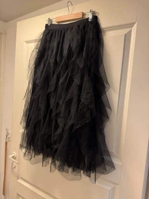 Svart volangkjol i tyll M - Säljer en svart långkjol i tyll med flera lager volanger som ger ett riktigt fluffigt och dramatiskt intryck. Kjolen har resår i midjan och är perfekt för dig som vill sticka ut med en edgy och cool look.