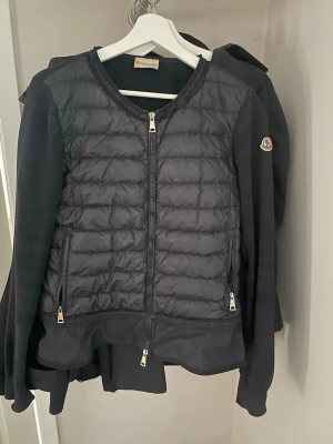 Marinblå quiltad carigan jacka från Moncler - Säljer en marinblå cardigan från Moncler med quiltad front och släta ärmar. Jackan har dragkedja framtill, två sidofickor med dragkedja och rund halsringning. Klassisk Moncler-logga på ärmen. Jackan har en volang nertill. Den är använd men riktigt bra sick, inga defekter alls. Jag har vanligtvis stl S och är 165 cm. 