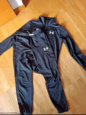 Under armour  - Bilden visar ett svart träningsset från Under Armour med byxor och jacka, men inga skor finns med.