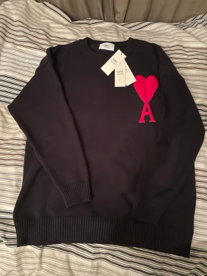 Svart stickad tröja från Ami - Svart stickad tröja från Ami Alexandre Mattiussi med röd hjärt- och A-detalj på bröstet. Klassisk rund halsringning och ribbade muddar vid ärmslut och nederkant. Perfekt för dig som gillar stilrena plagg med en twist.