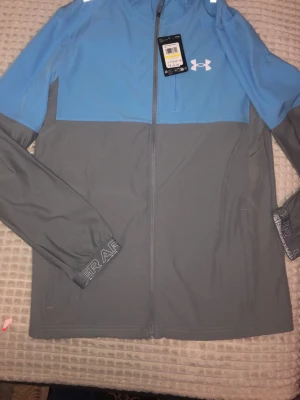Blå och grå vindjacka från Under Armour - Snygg tvåfärgad vindjacka från Under Armour i ljusblått och grått.  Är inte använd alls. Äkta med prislapp och allt kvar. Ni kan se på bilderna hur nya dem är. Kostar 1200 i butik men jag säljer dem endast för 800.
