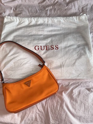 Orange axelväska från Guess - Säljer en snygg orange axelväska från Guess med klassisk trekantslogga i metall framtill. Väskan har justerbart axelband, dragkedja upptill och ljusbeige foder med Guess-tryck. Perfekt storlek för det viktigaste och ger en färgklick till din outfit. Bredd är 27 cm och höjd är 13 cm.