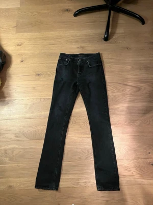 Svarta skinny jeans från Nudie Jeans - Lite schystare Svarta jeans från Nudie Jeans med klassisk femficksdesign och slitna detaljer framtill. Jeansen har en mörk tvätt och är tillverkade i stretchig bomullsdenim för en jätte bra passform. Snygga bakfickor med Nudie-logga och läderpatch i midjan.