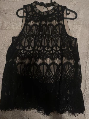Svart spetsblus  - Säljer en svart blus i spets med hög hals och ärmlös design. Blusen har ett vackert spetsmönster och transparent tyg med nude-färgad underdel. Perfekt för dig som gillar detaljer och vill sticka ut.