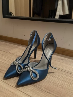 Blå pumps med strassrosett - Snygga blå pumps i satin från Catwalk med spetsig tå och hög klack. Skorna har en elegant rem runt vristen och är dekorerade med en glittrig strassrosett framtill. Perfekta för dig som vill sticka ut med lite extra bling.
