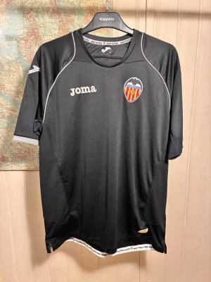 Valencia CF 2011/2012 Joma away shirt, size L - Good condition.  #valencia #valenciacf #laliga #spain #joma #soldado #banega #alba  Svart fotbollströja med Valencia CF-emblem och Joma-logotyp. Tröjan har grå detaljer på axlarna och ärmarna, samt en vit rand med text runt halsen.