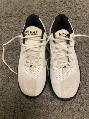 Nike G.T. Cut Academy vita sneakers - Nike G.T. Cut Academy sneakers i vitt med svarta detaljer och klassisk Swoosh på sidan. Skorna har snörning, meshpaneler för bra ventilation och en slitstark gummisula. Perfekta för dig som vill ha stilrena och sportiga skor till träning eller vardag.