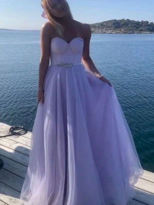 Ljuslila balklänning - Superfin ljuslila balklänning med axelbandslös överdel och markerad byst. Kjolen är lång och består av flera lager tyll som ger en riktigt prinsessig känsla. Midjan har ett smalt bälte med dekorativt spänne. Perfekt för bal eller student! Ingår gör då alltså bältet samt fodral till klänningen. Köpte den på Dressing Room Linné och har endast använt den en gång! Orginalpris 3200