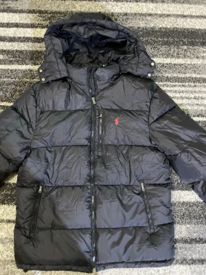 Svart pufferjacka från Polo Ralph Lauren - Säljer en svart pufferjacka från Polo Ralph Lauren med huva och dragkedjor. Jackan har det klassiska röda Polo-logot på bröstet och flera praktiska fickor med dragkedja. Perfekt för kalla dagar och riktigt snygg till streetwear-stilen.