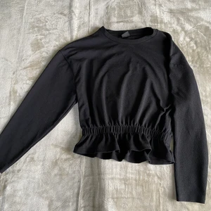 Tröja från Ginatricot - En svart långärmad tröja, med resår i midjan med en volang under. 