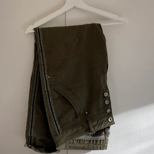 Cream denim militär byxor med revärer  - Byxor i militärgrönt med knappar fram och med fem fickor, två bak och tre fram. Svarta revärer på sidan och är i byxstorlek storlek 32 (midjemått) och något lägre i midjan är ”regular waist”. Style: Stine twill pants- Bailey fit. Nypris 1200kr 