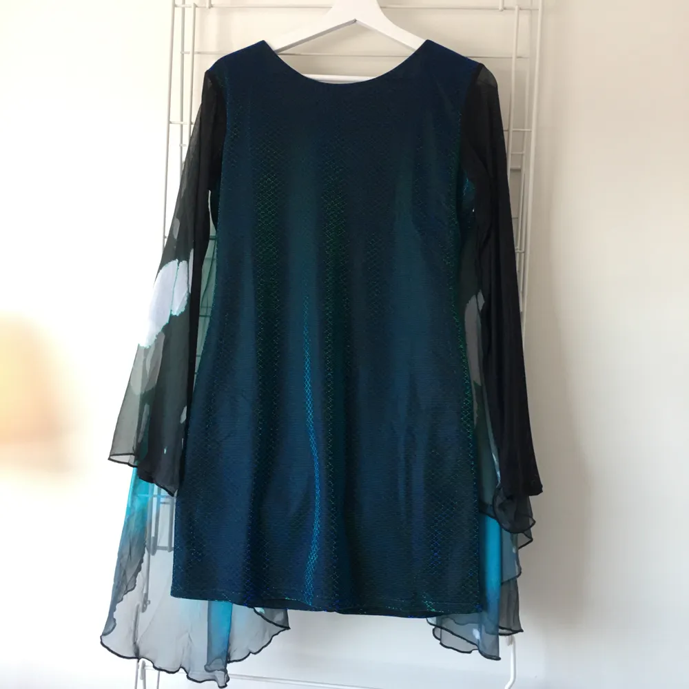 The label is cut off but it fits size M/L. The length from neck down is size 75cm. Dress has been used twice, and one of the arms has been a little damaged around the hand/wing, but it seems easy enough to sew together! The dress has a black colour with shimmering dark blue and dark green. The arms are half transparent and the wrists connect decorated butterly 
