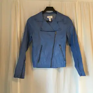 Blå jacka i fakeskinn från Issue 1.3. Två fickor med dragkedja framtill. Jackan stängs med både knäppning och dragkedja.