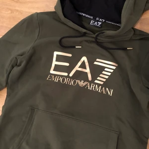 Emporio Armani hoodie - Militärgrön hoodie/tjocktröja från Emporio Armani med gulddetaljer och guldtryck. Mjukt material. Sparsamt använd. Säljer pga att jag har vuxit ur den. Storlek XS. Inköpt för 999kr, säljs nu för 199kr. Först till kvarn!