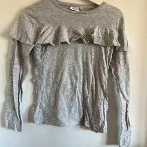 Grå tröja med volang  - En grå tröja med volang i storleken 158/164. Från Kappahl. Säljer då den är för liten. Säljer den för 60 kr och frakt ingår i priset så du får hela tröjan för 60 kr :)