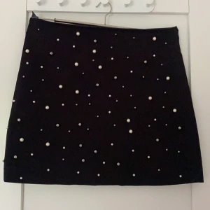 Snygg kjol med pärlor - Supersnygg svart kjol från New Yorker. På kjolens framsida finns pärlor fast sudda i olika storlekar samt på baksidan är det helt svart. Kjolen är i mocka material samt att det finns en dragkedja som man drar upp på baksidan (den syns knappt). Kjolen kort och i väldigt bra skick! 