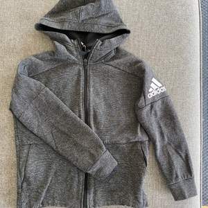 Svart/grå melerad Adidas hoodie med dragkedja. Storlek 140 (UK 9-10 år). Fickor i sidorna med dragkedjor.
