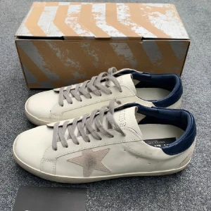 Vita sneakers med grå stjärna - Snygga vita sneakers från Golden Goose med grå mockastjärna på sidan, grå snörning och mörkblå häl. Skorna har rund tå och platt sula, tillverkade i skinn med detaljer i mocka. Perfekta för dig som gillar en clean men ändå unik look.