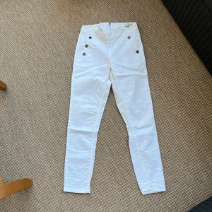 Vita  byxor från Newhouse - Snygga vita byxor från Newhouse med hög midja och smal passform. Byxorna har dekorativa knappar framtill och dragkedja bak. Tillverkade i stretchig bomull för en fräsch och stilren look. Perfekta till sommaren. Storlek 34.