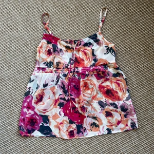 Blommigt linne från Esprit - Supersött linne från Esprit med blommigt mönster i rosa, orange och svart.Tunt, luftigt tyg med smala reglerbara axelband och knyt framtill. Perfekt för varma dagar när du vill ha en chill och fräsch look. Storlek 34. 