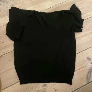 Svart blus med volangärm - S/M En svart blus med snygga volangärmar och rak passform. Toppen är enkel men har en cool detalj med de breda volangerna på axlarna som ger en trendig look. Perfekt att styla med jeans eller kjol för en stilren outfit.