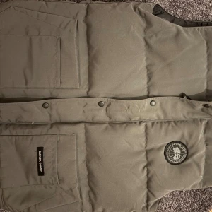Grå dunväst från Canada Goose  - Säljer en grå dunväst från Canada Goose i storlek XL men passar dom som har L Västen har två stora fickor framtill, tryckknappar och klassisk Canada Goose-logga på bröstet. Perfekt för lager-på-lager och håller dig varm under kyliga dagar 