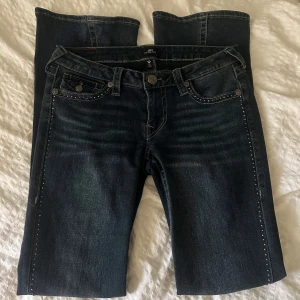 Mörkblå True Religion jeans med nitar - Snygga mörkblå jeans från True Religion med dekorativa nitar längs fickor och bensidor. Klassisk femficksmodell med markerade sömmar och logga på bakfickan. Jeansen har utsvängda ben och cool faded effekt framtill. Storlek 27 och passar mig som är 173cm. Nyskick! 