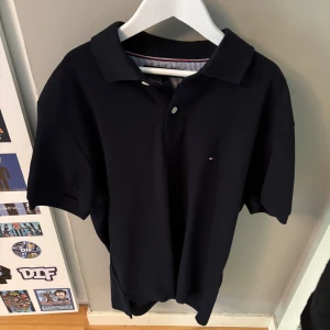 Svart pikétröja från Tommy Hilfiger - Klassisk svart pikétröja från Tommy Hilfiger med liten logga på bröstet. Tröjan har krage, korta ärmar och knäppning framtill. Perfekt för en clean och stilren look. Materialet är mjuk bomull som känns skönt mot huden.