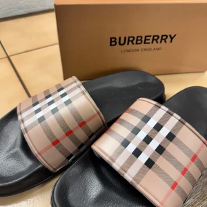 Burberry sandaler med klassiskt rutmönster - Snygga sandaler från Burberry med det ikoniska beige, svarta och röda rutmönstret på remmen. Svart sula i syntetmaterial ger en clean look och skön känsla. Perfekta för sommardagar och chill vibes. Med box