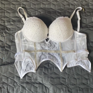 Vit korsett med spets och strassdetaljer - Supersnygg vit korsett med vadderade kupor, spetsdetaljer och glittrande strassband. Justerbara axelband och hakknäppning bak. Transparent mesh och dekorativa band ger en lyxig känsla. Perfekt för att lyfta vilken outfit som helst. Strassbandet har lossnat lite på ena bandet men går och limma 