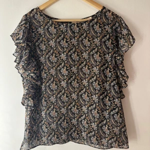 Blommig blus från Rue de Femme - Säljer en transparent blus från Rue de Femme med blommigt mönster i svart, beige och grått. Blusen har volangdetaljer på ärmarna och en luftig, flowy passform. Perfekt för dig som gillar unika prints och lätta material.