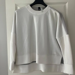 Vit sweatshirt från Adidas med dragkedja - Säljer en oversized vit sweatshirt från Adidas med grå panel på ryggen och axlarna. Tröjan har lång ärm, rund halsringning och en snygg dragkedjedetalj på axeln. Adidas-logga framtill.