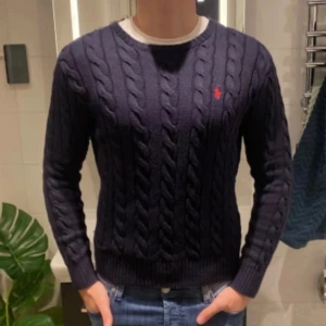 Ralph Lauren Kabelstickad tröja - Säljer en stilren mörkblå kabelstickad tröja från Polo Ralph Lauren med det klassiska röda logotypbroderiet på bröstet. Tröjan har en rund halsringning och långa ärmar, perfekt för alla årstider. Storlek S passar L. Kan tänka mig sänka priset något vid en snabb och smidig affär.